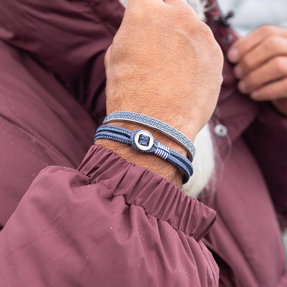 A close-up of a person's wrist wearing two braided bracelets: one dark blue and one light blue with a silver-toned square buckle, worn over a burgundy jacket.
