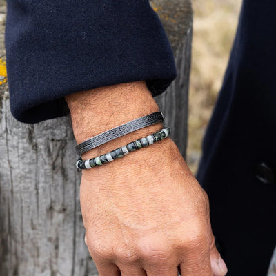A close-up of a person's wrist wearing two bracelets: a dark textured leather bracelet and a beaded bracelet with gray, white, and black stones, set against a rustic wooden post background.