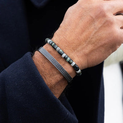 A close-up of a person's wrist wearing two bracelets: a dark gray engraved metal band and a beaded bracelet with green, white, and gray round beads.