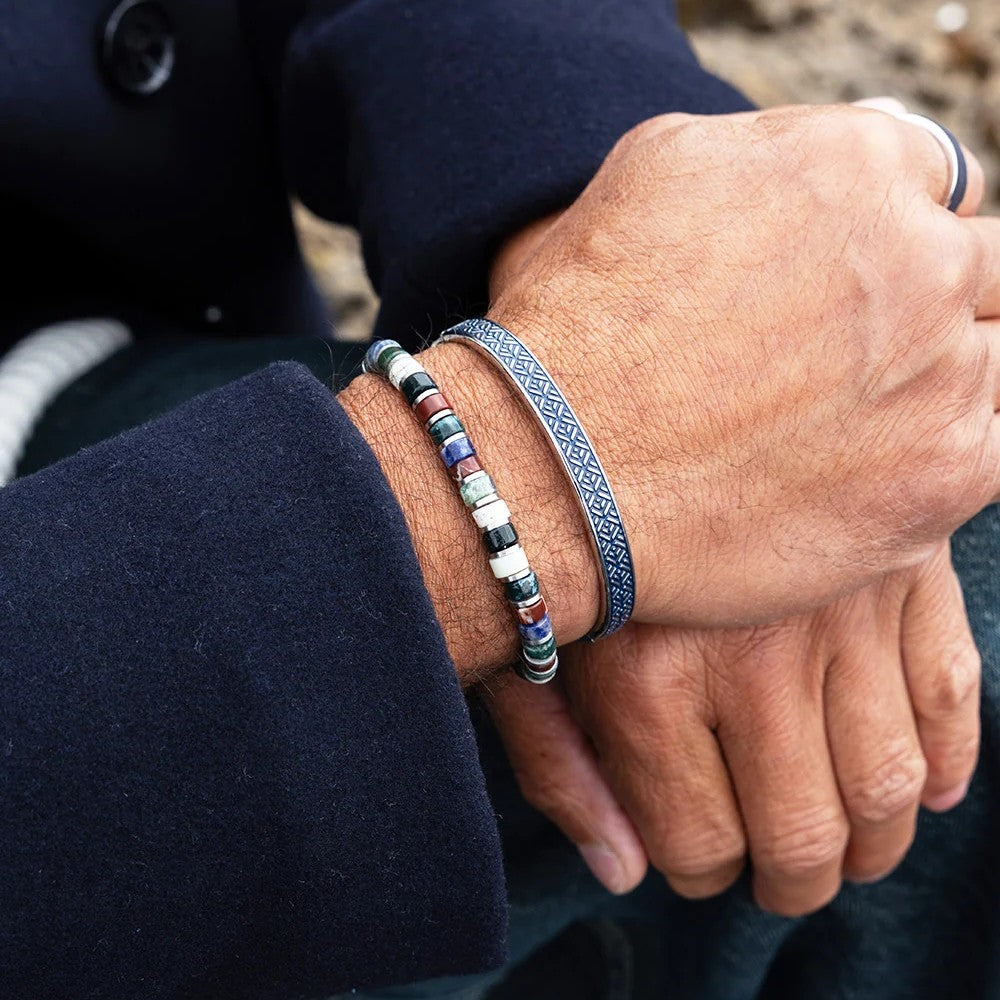 A close-up of a person's hands resting on their lap, wearing two bracelets: a silver-toned beaded bracelet with multicolored round beads and a silver-toned geometric patterned bracelet.