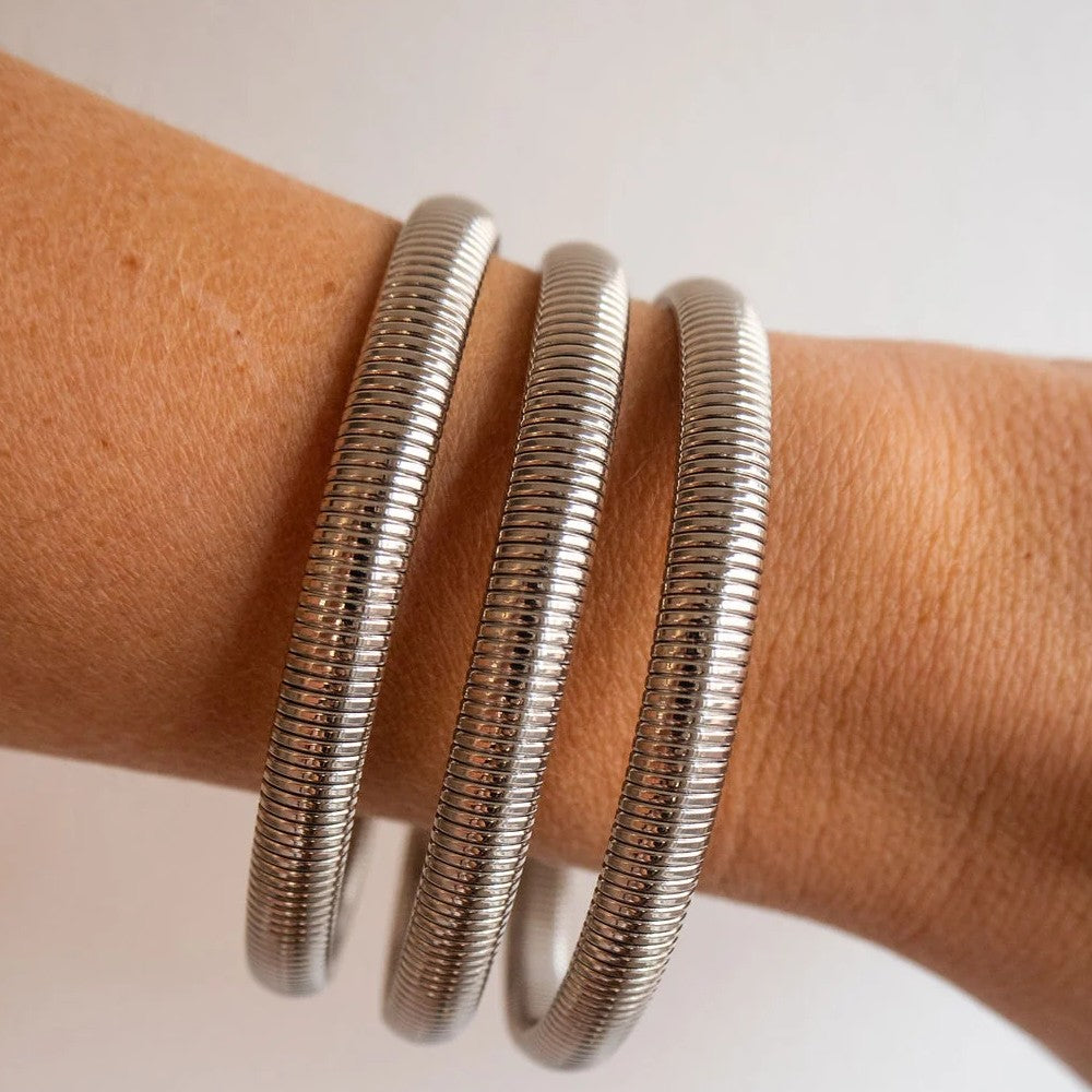 Three slim silver snake-style bangles wrapped on a person's wrist, shown against a plain light background.