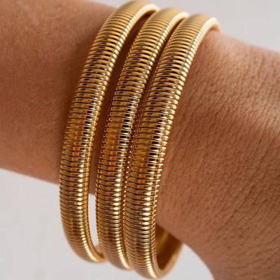 Three stacked gold snake-bangle bracelets worn on a person's wrist, shown in a close-up shot against a light neutral background.