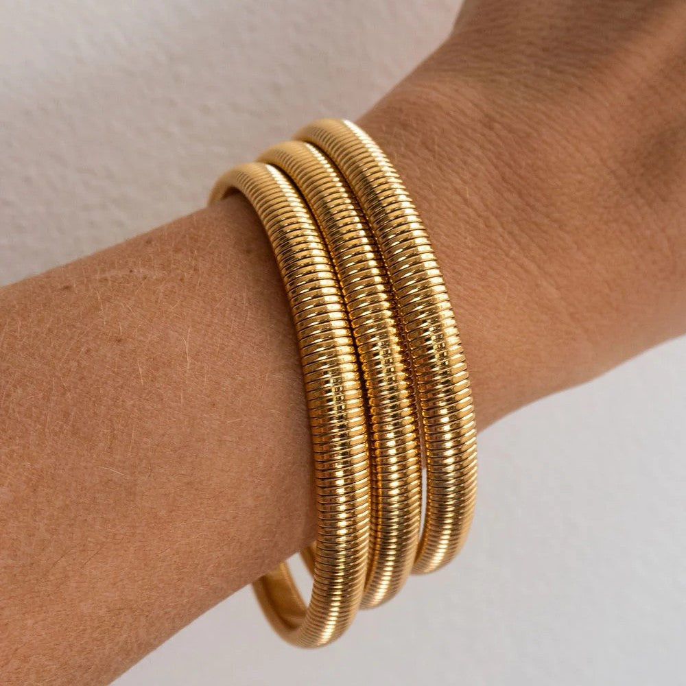 Three stacked gold snake-bangle bracelets worn on a person's wrist, shown against a plain light background.