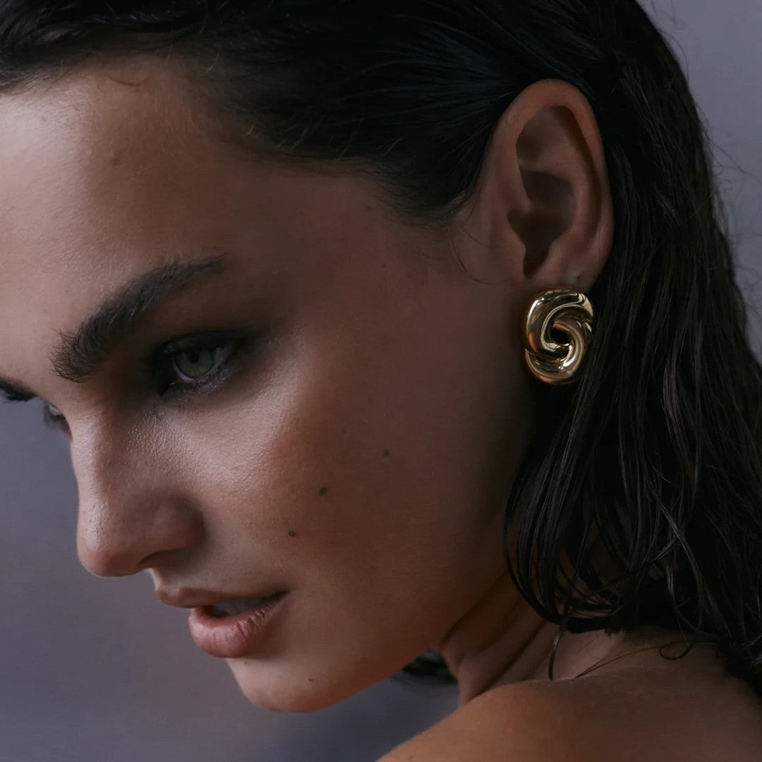 A close-up of a woman with dark hair wearing a gold spiral-shaped earring, shown in profile against a soft gray background.