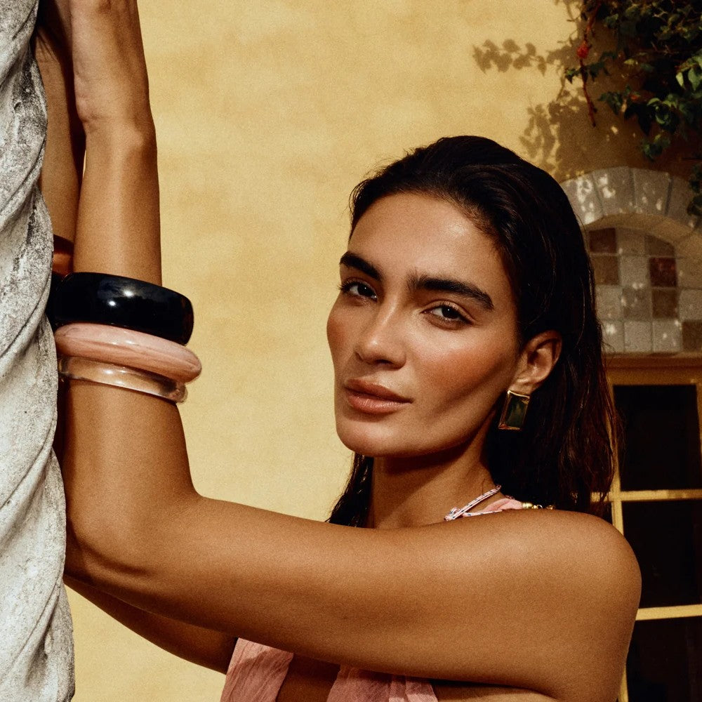 A woman wearing stacked bangle bracelets, including a prominent black bangle, a light pink bangle, and a clear silicone bangle, posing against a warm beige stucco wall.