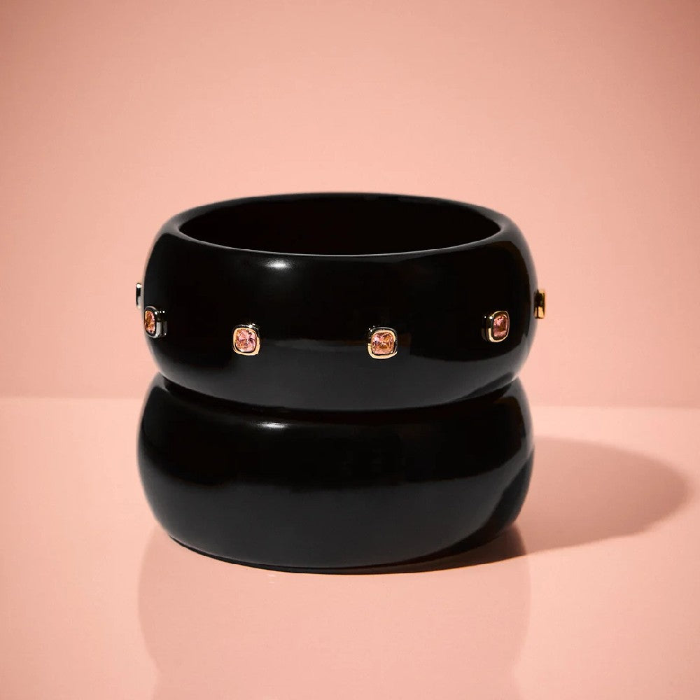 A glossy black stacked bangle bracelet with small gold-toned and pink gemstone accents, displayed on a soft pink reflective surface.