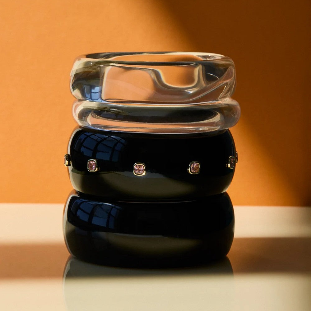 Three stacked bangles: a clear textured glass top bangle, a glossy black middle bangle with small pink gemstone accents, and a larger opaque black bottom bangle, set against a warm orange background.