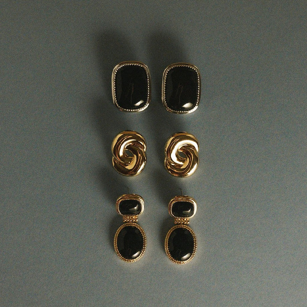 Three pairs of gold-tone earrings arranged vertically on a dark gray background. The top pair has rectangular black stones, the middle pair has twisted gold-tone loops, and the bottom pair has double stacked rectangular black stones with gold-tone borders.
