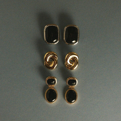 Three pairs of gold-tone earrings arranged vertically on a dark gray background. The top pair has rectangular black stones, the middle pair has twisted gold-tone loops, and the bottom pair has double stacked rectangular black stones with gold-tone borders.