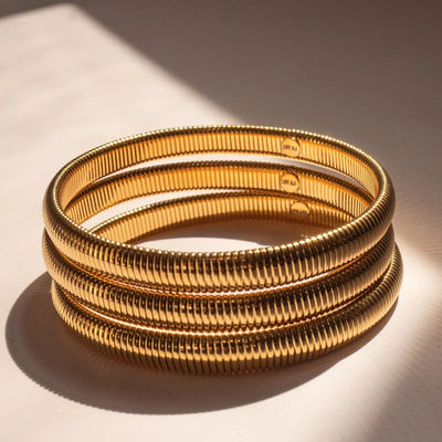 Three stacked gold-toned bangles with a ribbed, snake-like textured pattern, photographed on a warm beige surface with dramatic diagonal sunlight and shadows.