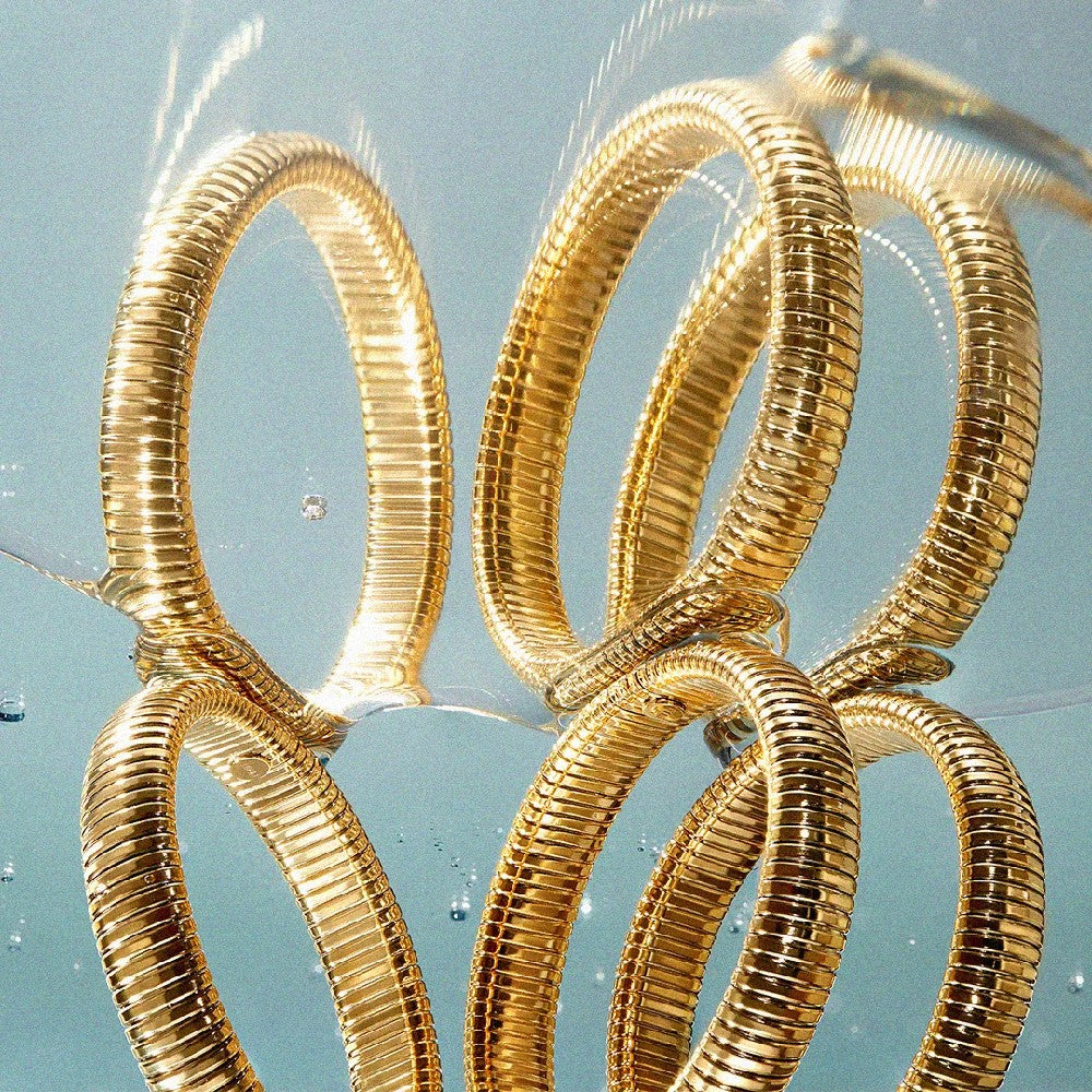 A close-up of several interlocking metallic rings with a ribbed, coiled texture in a shiny gold finish, set against a pale blue background with soft reflections and small bubbles.