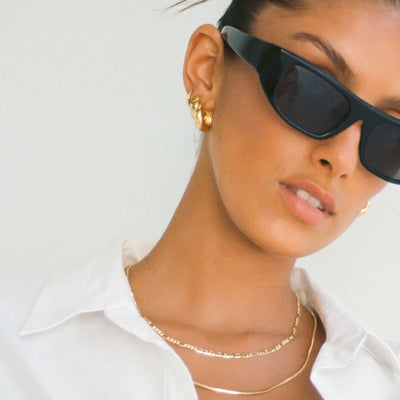 A close-up lifestyle shot of a model wearing black cat-eye sunglasses, gold hoop earrings, and layered gold necklaces, wearing a white collared shirt against a plain white background.