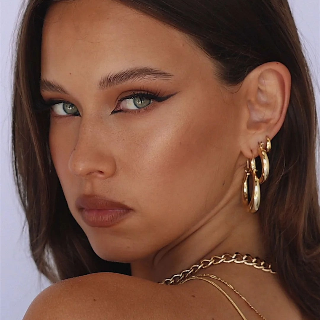 A close-up portrait of a woman wearing large gold hoop earrings and layered gold necklaces, with winged black eyeliner and a neutral expression against a light background.