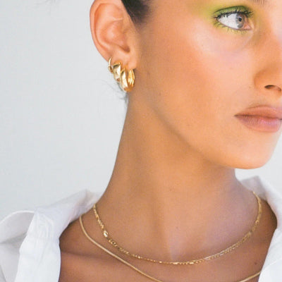A close-up lifestyle shot of a model wearing a pair of gold twisted hoop earrings, a delicate gold chain necklace, and a thicker gold chain necklace, paired with a white collared shirt against a light gray background.