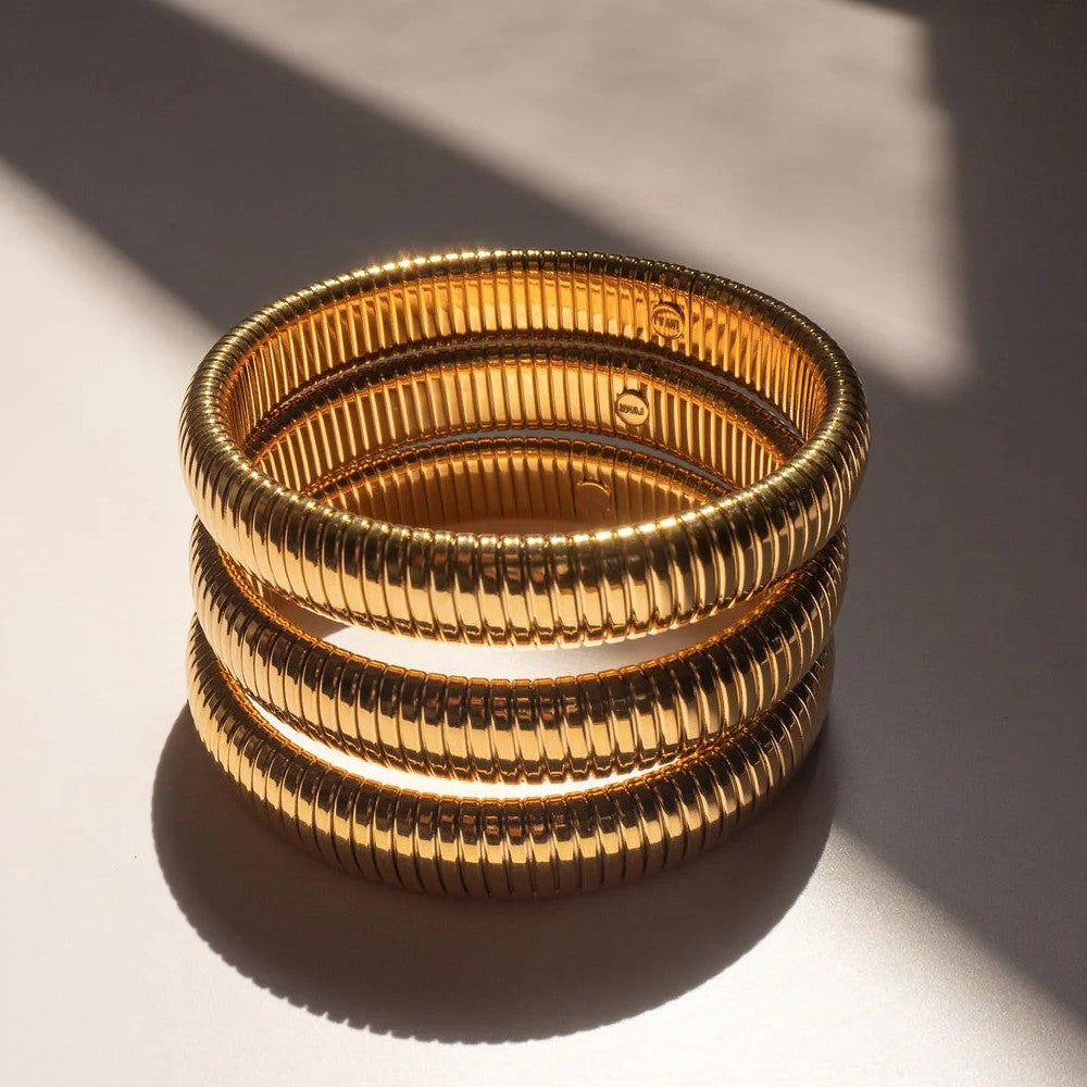 Three stacked gold-toned ribbed bangles with a textured, snake-like exterior, photographed on a white surface with dramatic diagonal shadows.