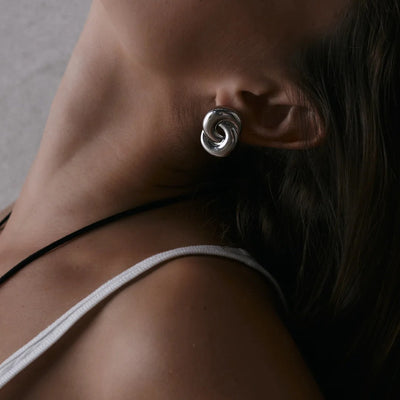 A close-up of a woman's ear wearing a polished silver spiral-shaped stud earring, with her neck, shoulder, and dark hair visible against a neutral gray background.