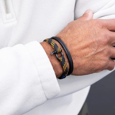 A close-up of a person's wrist wearing two bracelets, including a black and yellow braided cord bracelet with a toggle clasp and black leather band, paired with a white zip-up sweatshirt.
