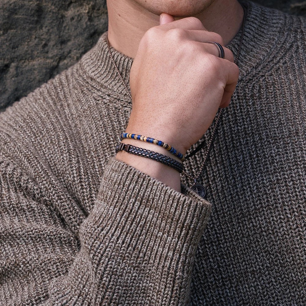 Close-up of a person wearing three beaded and braided bracelets on the wrist, with a textured brown knit sweater and a dark stone background.