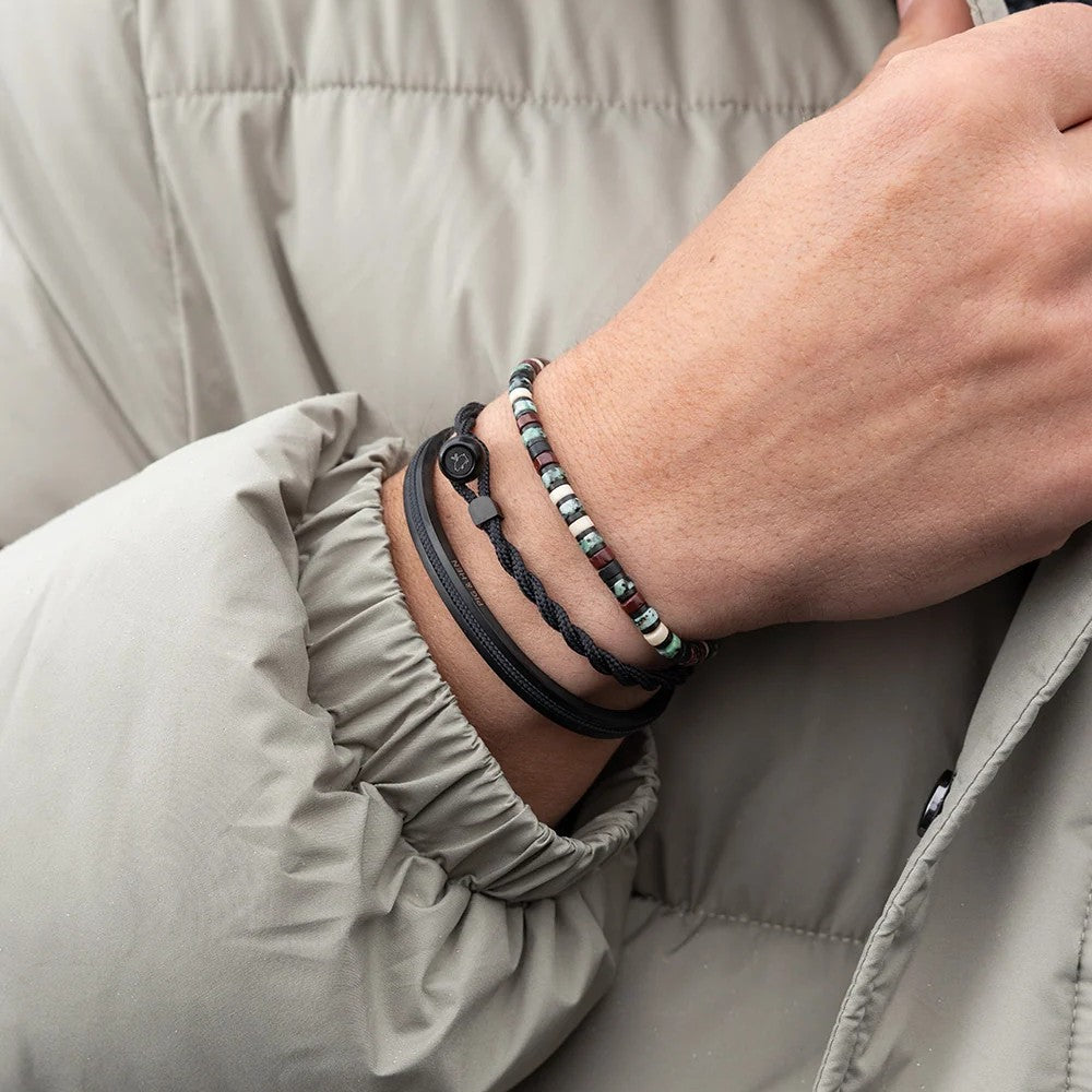 A close-up of a person's wrist wearing multiple bracelets, including a black braided cord bracelet, a black adjustable cord bracelet with a toggle clasp, and a beaded bracelet with small multicolored beads, all worn over a light beige puffer jacket.
