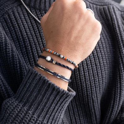A close-up of a person's wrist wearing multiple bracelets, including a beaded bracelet with orange and gray beads, a dark braided cord bracelet, a silver-toned bangle, and a thin black bracelet, worn over a dark gray ribbed sweater.