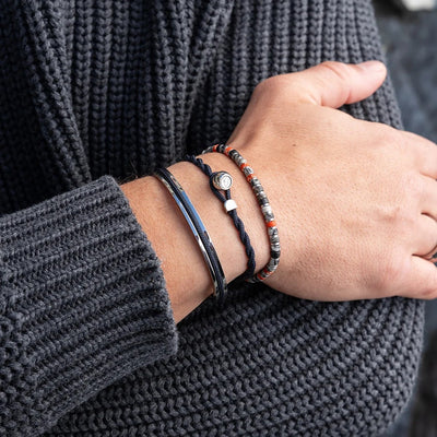 A close-up of a person's wrist wearing three distinct bracelets, including a beaded bracelet with red, gray, and white beads, a black cord bracelet with a silver charm, and a dark blue cord bracelet, all worn over a dark gray knit sweater.