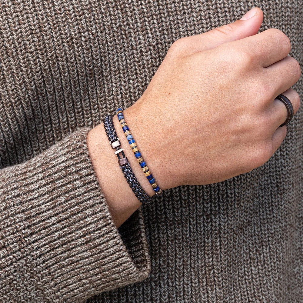 A close-up of a person's hand and wrist wearing two beaded bracelets against a brown knit sweater. One bracelet is dark gray with small metallic beads, and the other is blue and gold with metallic spacer beads.