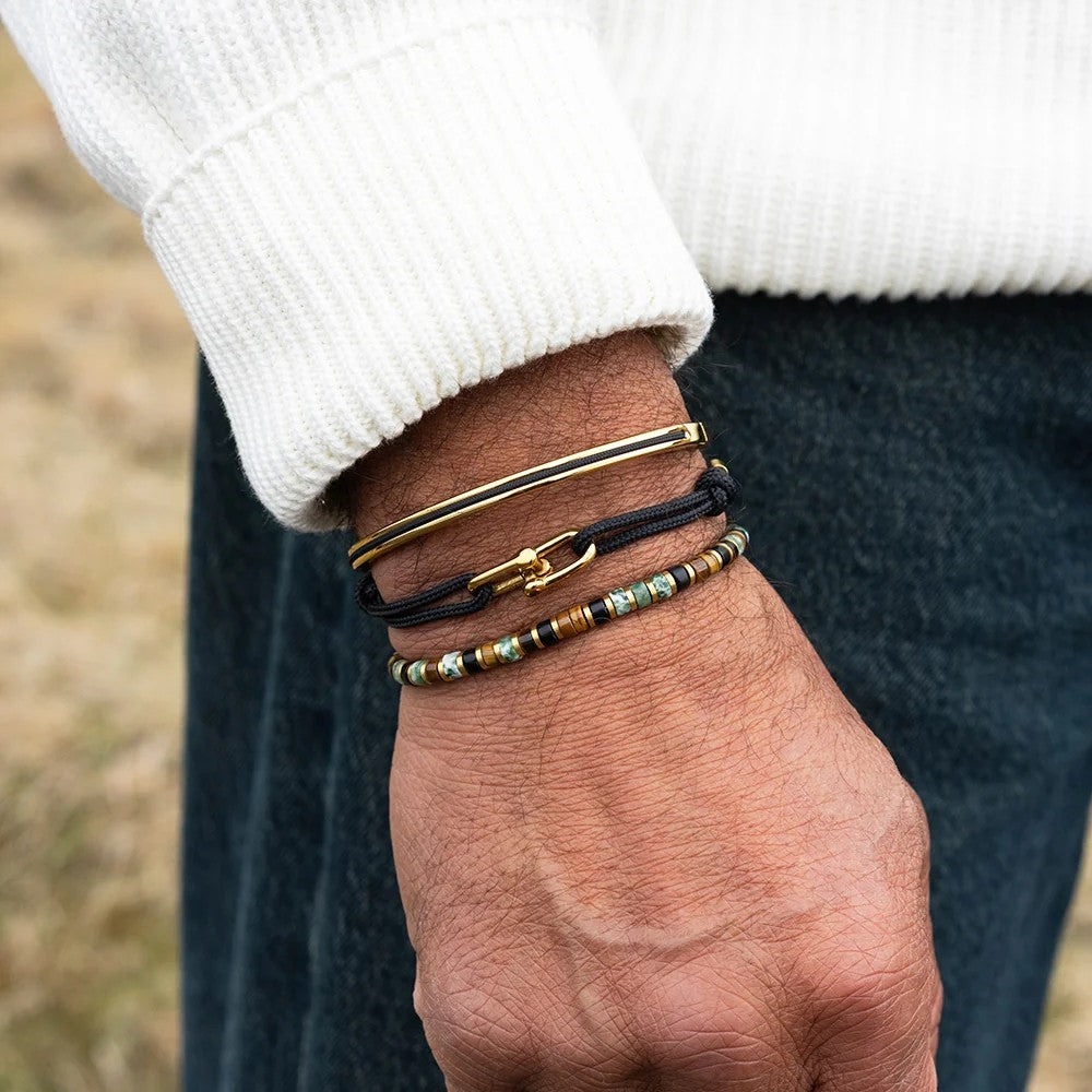 A close-up of a person's wrist wearing three stacked bracelets: a gold bangle, a black cord bracelet with a gold clasp, and a black cord bracelet with multicolor beads.