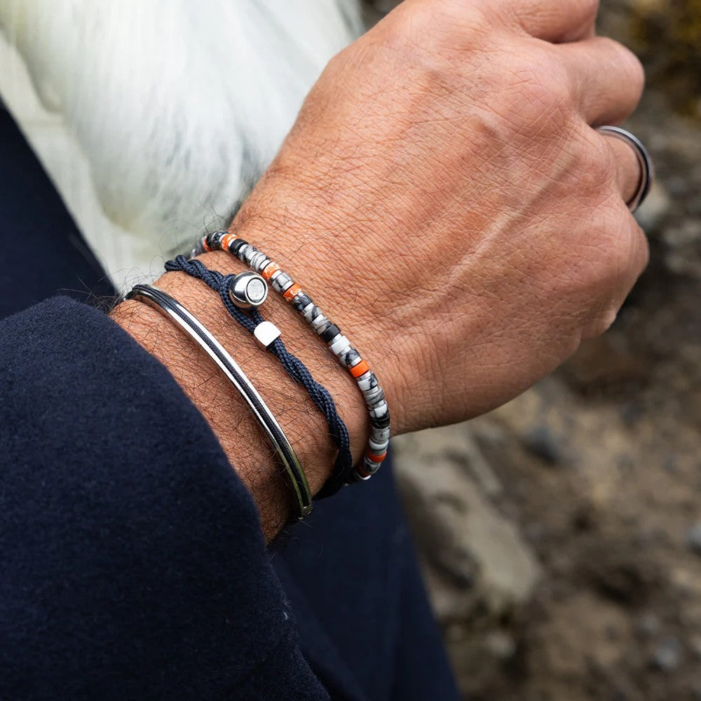 A close-up of a person's wrist wearing multiple bracelets, including a dark navy braided cord bracelet with a silver-toned oval charm, a silver bangle, and a beaded bracelet with gray, white, and orange beads. The background is a soft-focus outdoor scene.