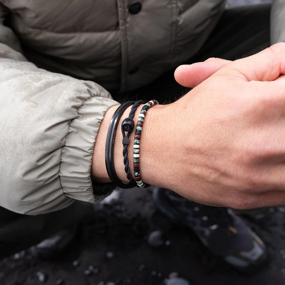 A close-up of a person's wrist wearing multiple bracelets, including a black cord bracelet with a silver bead, a braided black cord bracelet, and a beaded bracelet with silver, red, and white beads. The person is wearing a gray puffer jacket, and wet dark rocks are visible in the background.