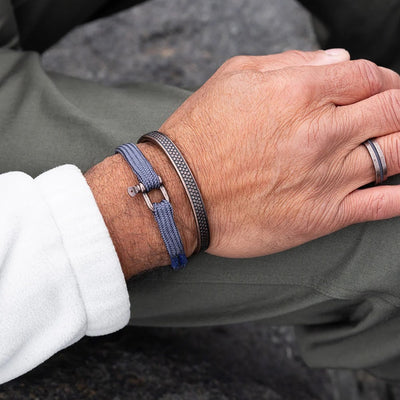A close-up of a person's wrist wearing two bracelets: a textured silver-toned metal bangle and a blue braided cord bracelet with a silver toggle clasp. The person is also wearing a silver ring on their finger, and the background shows olive green pants and a dark outdoor surface.