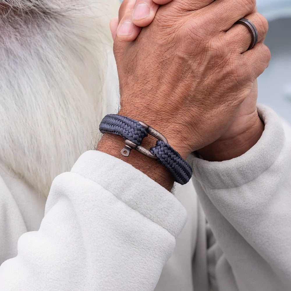 A close-up of a person's hands clasped together, wearing a dark gray braided rope bracelet with a silver metal clasp. The person is wearing a light gray fleece jacket.