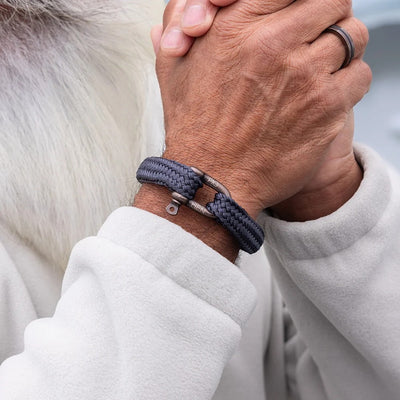 A close-up of a person's hands clasped together, wearing a dark gray braided rope bracelet with a silver metal clasp. The person is wearing a light gray fleece jacket.