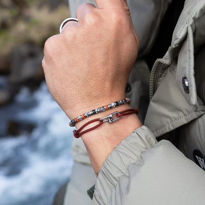 A close-up of a person's wrist wearing two bracelets: one beaded bracelet with red, gray, and white beads, and one dark red cord bracelet with a silver clasp. The person is wearing a light gray jacket, with a blurred outdoor background showing water and rocks.