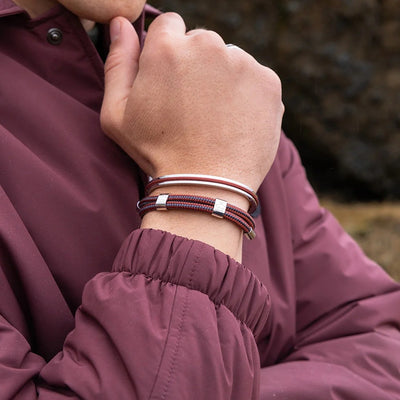 A close-up of a person wearing a stack of three bracelets on their wrist, including two burgundy cord bracelets with silver-toned square links and one silver-toned metal band. The person is wearing a matching burgundy jacket, with a blurred outdoor background.