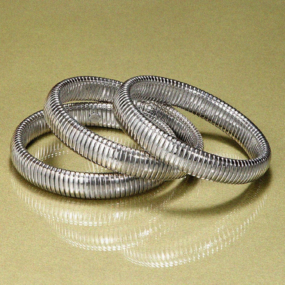 Three silver metallic bangles with a ribbed, textured design, arranged in a knot on a reflective gold surface.
