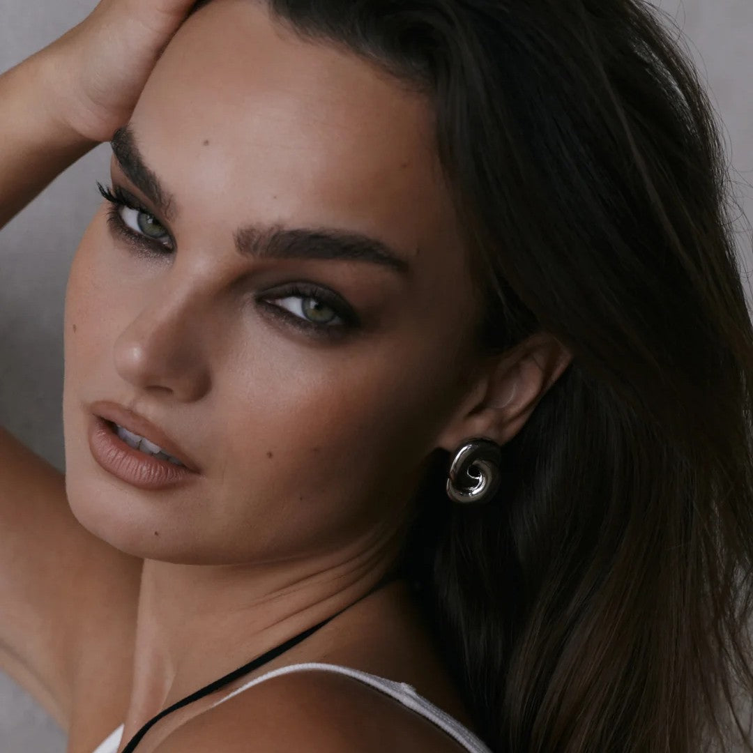 A close-up portrait of a woman with dark hair wearing a silver spiral-shaped hoop earring, looking directly at the camera.