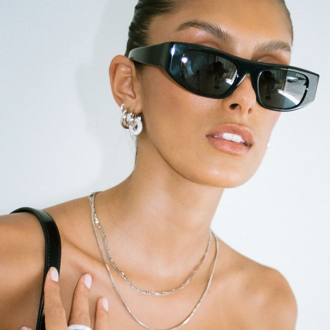 A close-up of a model wearing black cat-eye sunglasses, silver layered necklaces, and silver hoop earrings against a plain white background.