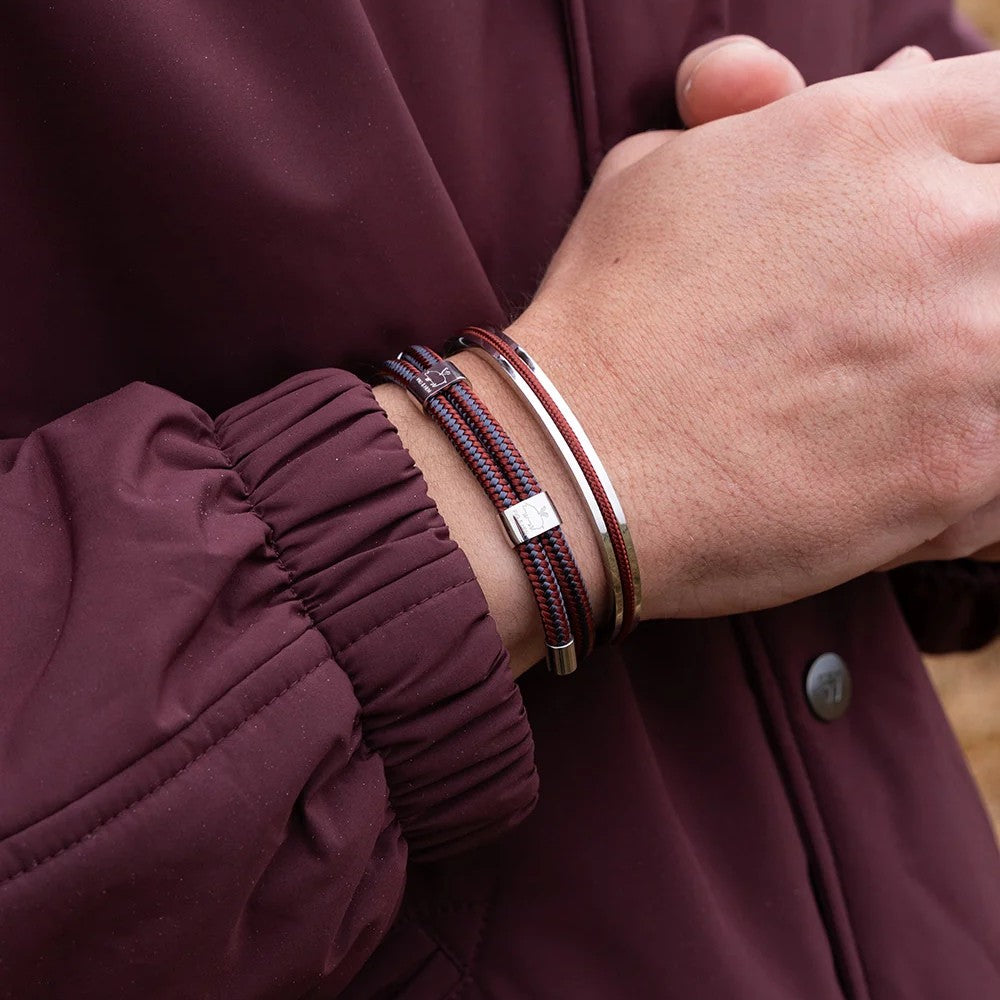 A close-up of a person's wrist wearing three bracelets: two dark red and black braided cords with silver square accents, and one slim silver metal band, all worn under a burgundy jacket.